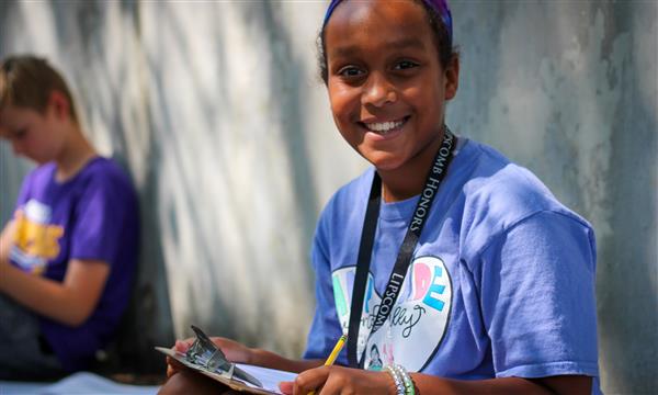 student smiles at camera while writing on clipboard